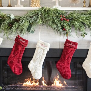 May include: Five knitted Christmas stockings hang above a fireplace. The stockings are white and red with a cable knit pattern. The stockings are personalized with the names "Dad", "Anna", "Joseph", "Mom", and "Grace".