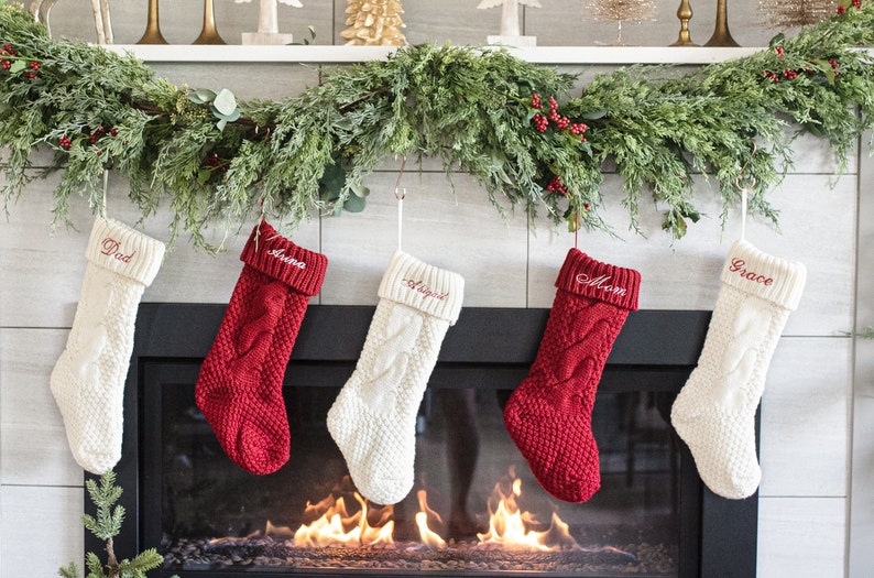 May include: Five knitted Christmas stockings hang above a fireplace. The stockings are white and red with a cable knit pattern. The stockings are personalized with the names "Dad", "Anna", "Joseph", "Mom", and "Grace".