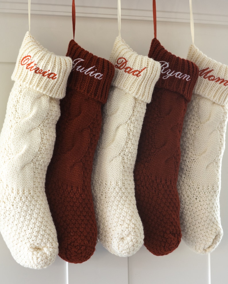 May include: Five knitted Christmas stockings with names embroidered on the cuff. The stockings are white and brown with a cable knit pattern. The names are Olivia, Julia, Dad, Ryan, and Mom.