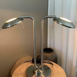 Könnte beinhalten: Eine Schreibtischlampe aus gebürstetem Metall mit zwei gebogenen Armen, die jeweils ein kreisförmiges Licht tragen. Die Lampe hat einen runden Sockel und steht auf einem Holztisch. Das Design ist modern und minimalistisch.