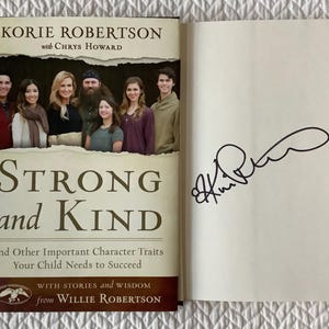 May include: A hardcover book titled Strong and Kind by Korie Robertson with Chrys Howard, featuring a photograph of eight people on the dust jacket. The open book displays a black ink signature on the blank white endpaper opposite the cover.