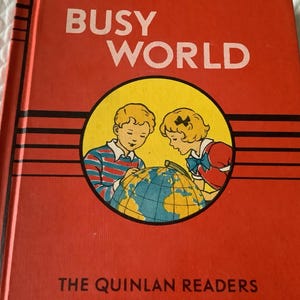 May include: A red book with the title "BUSY WORLD" and the text "THE QUINLAN READERS" on the cover. The cover features an illustration of two children looking at a globe.