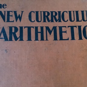 May include: The cover of an old book titled "The New Curriculum Arithmetics". The cover is brown with black text.