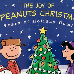 May include: A book cover featuring Charlie Brown and Lucy from the Peanuts comic strip, standing near a decorated Christmas tree. The title reads "The Joy of A Peanuts Christmas" with the subtitle "50 Years of Holiday Comics!" against a blue starry background.