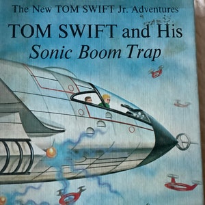 May include: The New Tom Swift Jr. Adventures book cover featuring a white and grey futuristic aircraft with two figures in the cockpit. The title reads "TOM SWIFT and His Sonic Boom Trap".