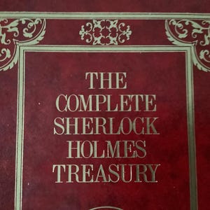 May include: A close-up of a book cover with the title "THE COMPLETE SHERLOCK HOLMES TREASURY" in gold lettering. The cover is a deep red with gold decorative borders and accents.