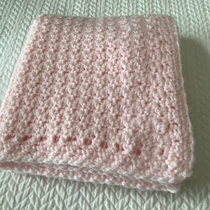 May include: A folded, light pink crocheted baby blanket. The blanket has a textured stitch pattern and a decorative border with small openings. The blanket is folded neatly on a white surface.