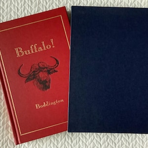 May include: A red hardcover book with a gold border and the title "Buffalo!" in gold lettering. The cover features a black and white illustration of a buffalo head. The author's name, "Boddington", is printed in gold lettering below the illustration.