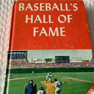 May include: A red book with the title "Baseball's Hall of Fame" in white text. The cover features a colorful illustration of a baseball game in progress, with players on the field and spectators in the stands. The text "Buy U.S. Bonds" is visible on a sign in the background.
