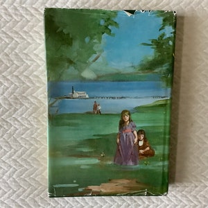May include: A vintage book cover with a green, blue, and brown illustration of two girls standing in a grassy field. The girls are wearing dresses and are looking at the viewer. In the background, there is a lake and a boat.