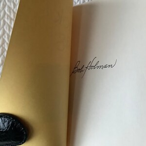 Op de afbeelding: Een wit boek met een gouden omslag is open op een blanco pagina. De pagina is gesigneerd met "Bob Holman" in zwarte inkt.