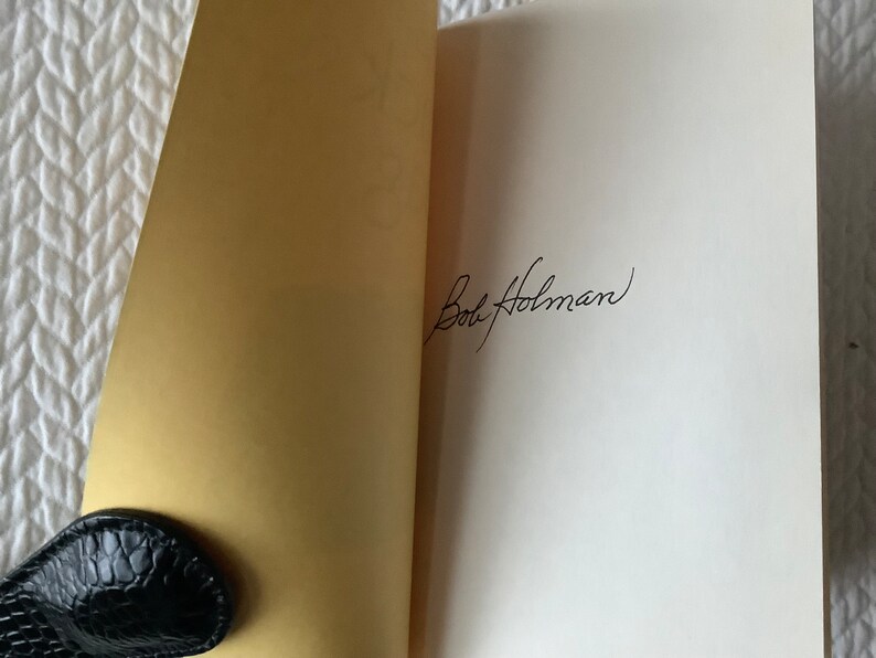 Op de afbeelding: Een wit boek met een gouden omslag is open op een blanco pagina. De pagina is gesigneerd met "Bob Holman" in zwarte inkt.