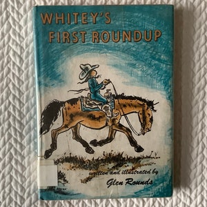 May include: A vintage book cover with a blue background and the title "Whitey's First Roundup" in yellow lettering. The cover features an illustration of a young person riding a horse.