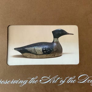 May include: A vintage duck decoy, painted in shades of black, brown, and white, rests on a cream-colored background. The decoy is displayed within a rectangular frame, with the text "Preserving the Art of the Decoy" in elegant script at the bottom.