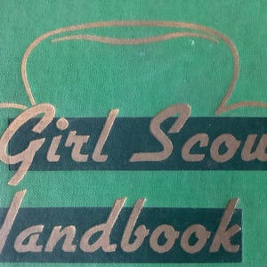 May include: A green book with a gold outline of a Girl Scout hat and the text "Girl Scout Handbook" in gold lettering.