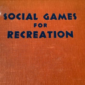 May include: An orange book cover with the title "SOCIAL GAMES FOR RECREATION" in dark blue lettering. The book appears to be vintage, with a textured surface and slight wear.