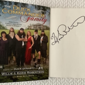 May include: A hardcover book titled The Duck Commander Family featuring a group photograph of six people on the cover, with the text How Faith, Family, and Ducks Built a Dynasty and Stars of Duck Dynasty on A&E. The adjacent page features a black ink signature.