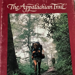 May include: A book cover with the title "The Appalachian Trail" in cursive script. The cover features a photograph of a person hiking through a lush, green forest with a backpack.
