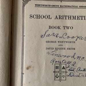 May include: Open book titled "School Arithmetics Book Two" by Sara Cooper, George Wentworth, and David Eugene Smith. The cover features decorative borders and handwritten inscriptions including "Linwood, n.c. Route #2."