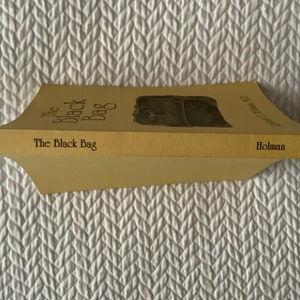 Op de afbeelding: Een geel paperback boek getiteld "The Black Bag" met een zwart-witte illustratie van een tas op de omslag. Het boek is uitgegeven door Holman.