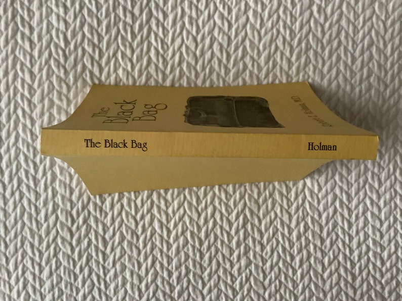 Op de afbeelding: Een geel paperback boek getiteld "The Black Bag" met een zwart-witte illustratie van een tas op de omslag. Het boek is uitgegeven door Holman.