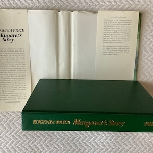May include: A green hardcover book titled "Eugenia Price Margaret's Story" with gold lettering. The book is closed and the spine is visible. The book is on a white surface.