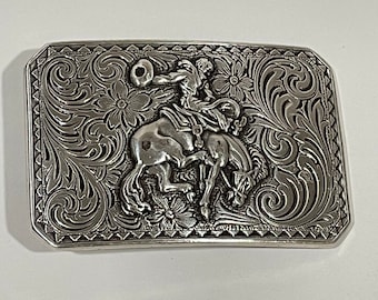 May include: Silver belt buckle with an intricate design featuring a cowboy riding a bucking bronco. The buckle has a detailed floral and scroll pattern.