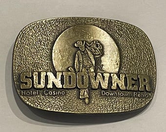 May include: A gold-toned metal belt buckle with a raised image of a cowboy in front of a sun. The buckle says "SUNDOWNER Hotel Casino Downtown Reno".