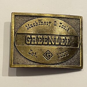 May include: A gold-toned metal belt buckle with the text "Machinery & Tools Greenlee Est. 1862" and a diamond-shaped "G" logo.