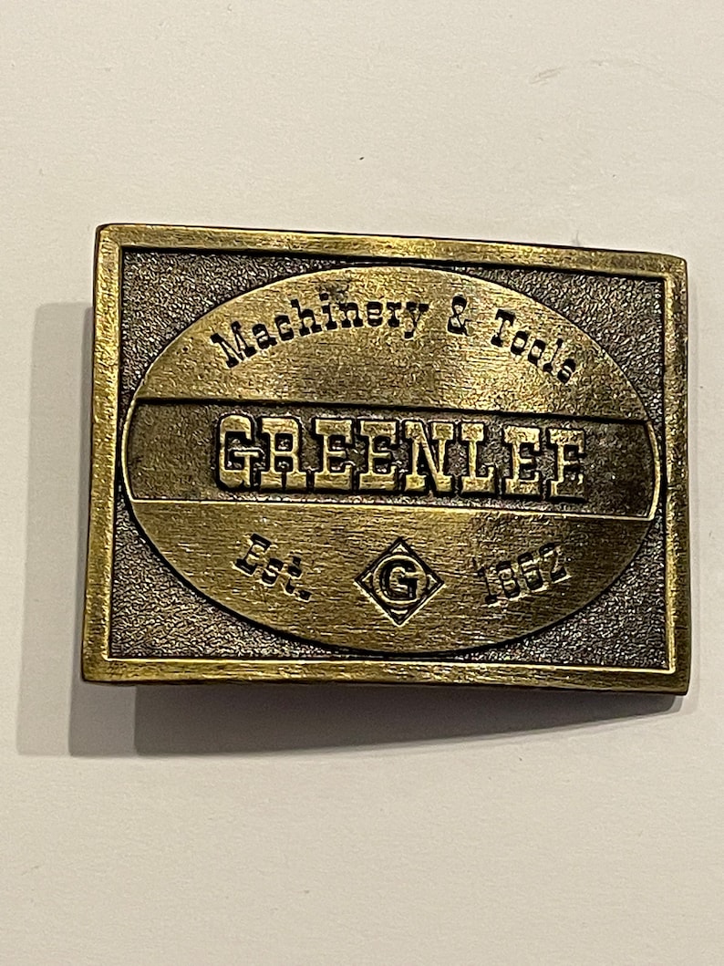May include: A gold-toned metal belt buckle with the text "Machinery & Tools Greenlee Est. 1862" and a diamond-shaped "G" logo.
