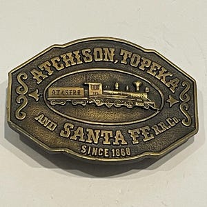 May include: A gold-colored metal belt buckle featuring a vintage train engine with the text "AT&SERR 225" on the side. The buckle also has the text "ATCHISON, TOPEKA AND SANTA FE R.R. CO. SINCE 1866".