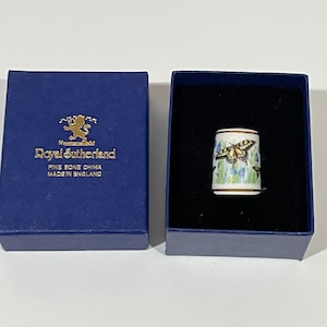 May include: A blue box with the text "Royal Sutherland Fine Bone China Made in England" and a lion logo. The box contains a white porcelain thimble with a gold trim and a butterfly design.