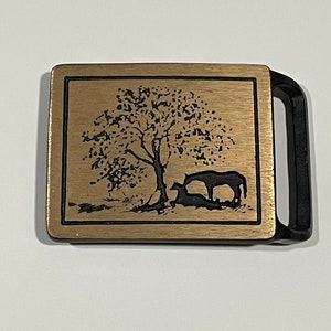 May include: A gold-toned belt buckle with a black silhouette of a tree and a horse. The buckle is rectangular with a smooth, brushed finish.