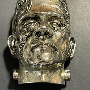 May include: A metallic silver-toned sculpture of Frankenstein's monster head. The piece features a flat top, prominent brow, textured hair, and two cylindrical bolts protruding from the sides of the neck. The surface has a reflective finish.
