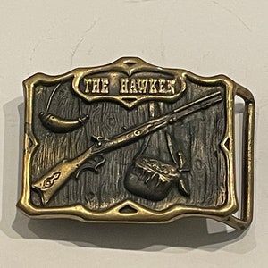 May include: A brass belt buckle with a detailed design of a rifle, a powder horn, and a horn. The buckle is engraved with the words "The Hawken".