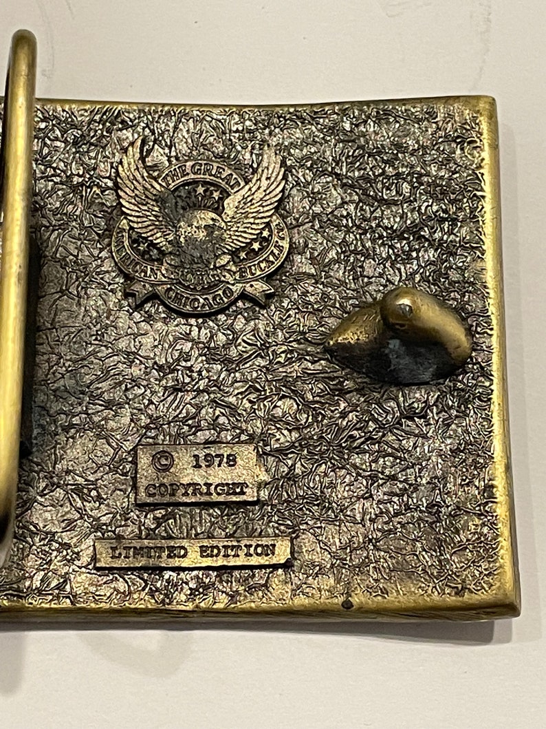 May include: A gold-toned belt buckle with a textured surface. The buckle features an eagle with the text "The Great American Solid Buckle Chicago" and the text "1978 Copyright Limited Edition".
