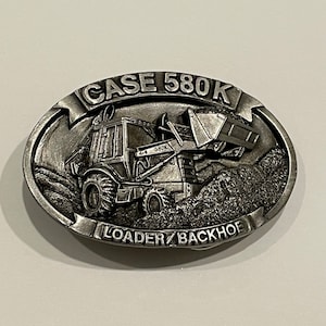 May include: A silver metal belt buckle featuring a Case 580K loader/backhoe with a detailed design. The buckle is oval shaped and has the words "CASE 580K LOADER/BACKHOE" engraved on it.