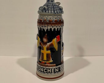 May include: A ceramic beer stein with a blue and gray roof, a black and yellow figure, and the word "MUNCHEN" on the bottom.