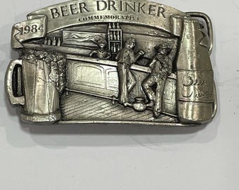 May include: A silver-toned belt buckle with a bar scene design. The buckle features the text "BEER DRINKER COMMEMORATIVE 1984". The design includes a beer mug, a beer bottle, and figures at a bar.