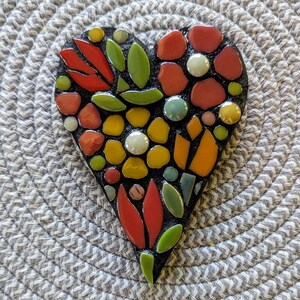 May include: A heart-shaped mosaic wall hanging featuring a black base decorated with various ceramic pieces in shades of red, yellow, orange, and green. The design includes floral and leaf patterns created with smooth, glossy tiles and round beads.