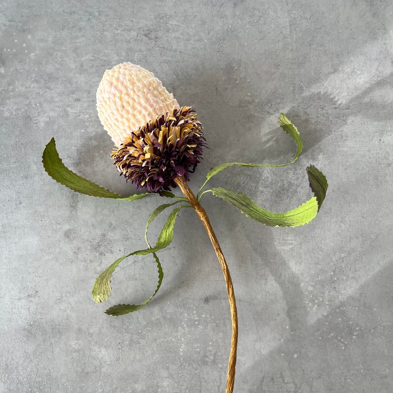 May include: A handmade Banksia flower featuring a cream-coloured, textured bloom, with a dark purple and yellow centre. The stem is brown, and the leaves are green and wavy. The flower is set against a grey background.