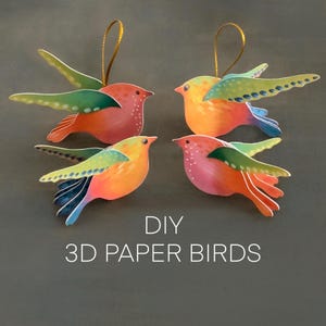 May include: Four colorful 3D paper bird ornaments with gold hanging strings. The birds have orange, pink, and yellow bodies with green and blue wings. The text "DIY 3D PAPER BIRDS" is below the birds.