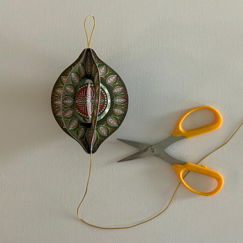 May include: An ornate, green and red paper ornament with a gold string loop, next to a pair of yellow-handled scissors. The ornament has a detailed floral design and is partially open, revealing its interior structure. The scissors are open, with the gold string running through them.