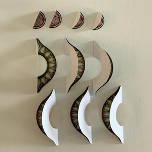 May include: Decorative wall art featuring a series of paper cutouts in a crescent moon shape. The cutouts have a white interior and a patterned exterior with red, green, and gold details. The art is arranged in a visually appealing pattern.