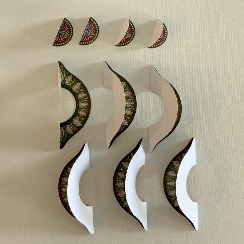 May include: Decorative wall art featuring a series of paper cutouts in a crescent moon shape. The cutouts have a white interior and a patterned exterior with red, green, and gold details. The art is arranged in a visually appealing pattern.