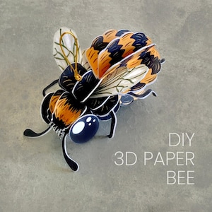 May include: A 3D paper bee craft featuring black and orange striped segments, translucent wings with green vein details, and large dark blue eyes. A gold hanging loop is attached to the top. The text DIY 3D PAPER BEE is printed in white.