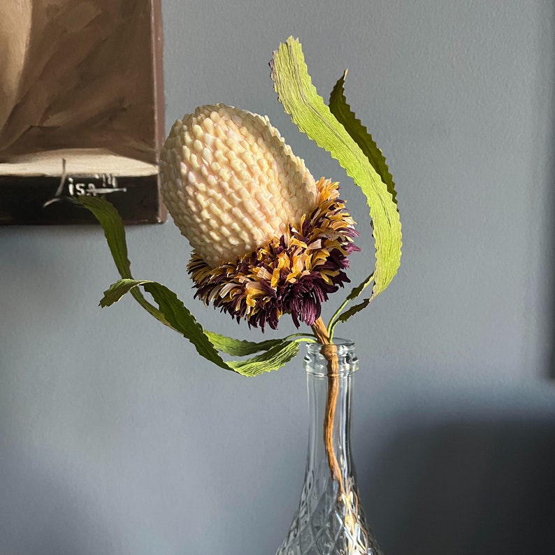 May include: A Banksia flower with a cream-coloured, textured head, and purple and yellow accents. The flower has green, wavy leaves and is displayed in a clear glass bottle. The background is a muted grey.