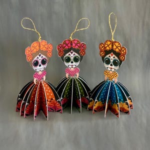 May include: Three hanging ornaments shaped like figures with sugar skull face paint and floral headpieces. Each figure wears a patterned bodice and a pleated, multi-colored skirt with floral motifs. Gold strings are attached to the top for hanging.