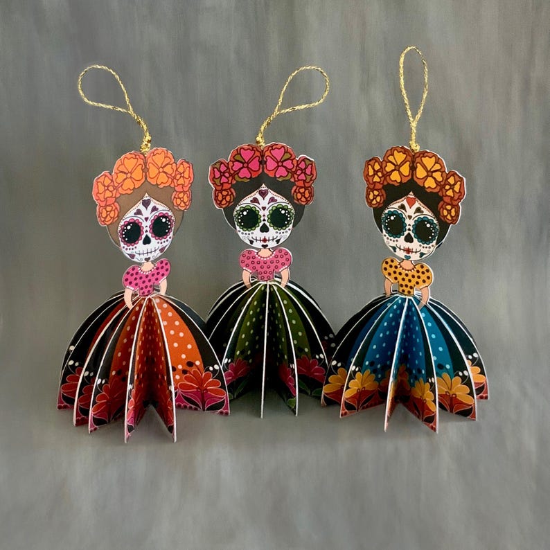 May include: Three hanging ornaments shaped like figures with sugar skull face paint and floral headpieces. Each figure wears a patterned bodice and a pleated, multi-colored skirt with floral motifs. Gold strings are attached to the top for hanging.