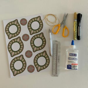 May include: An assortment of craft supplies including a sheet of decorative stickers with green and pink floral designs, scissors, pens, a ruler, clear glue, and gold string. The items are arranged on a white surface.